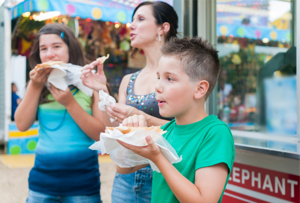 Three people enjoy funnel cakes at a vibrant fair with colorful booths, creating a fun atmosphere near Southport rentals.