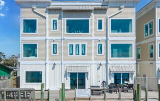 Three-story beige waterfront building with large windows and a wooden dock, ideal for Southport rentals. Sunny blue sky and some trees visible.