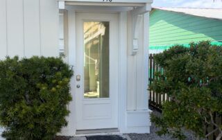 White door with small awning marked "140," flanked by shrubs, pathway leading to entry; Southport rentals ambiance.
