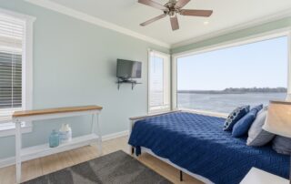Bright bedroom with blue bedding, large window with water view, ceiling fan, TV, and a white shelf. Ideal for Southport rentals.
