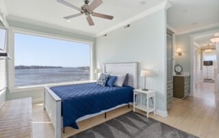 Cozy bedroom with blue bedding, large window overlooking water, wood floors, ceiling fan, ideal for Southport rentals.