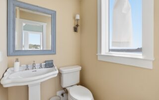 Bathroom with pedestal sink, toilet, and wall sconce. Large mirror and window with coastal view, highlighting Southport rentals ambiance.