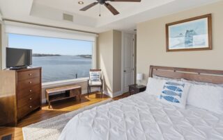 Cozy bedroom with large window overlooking water view. Features white bedding, wooden furniture, TV, and nautical decor. Ideal for Southport rentals.