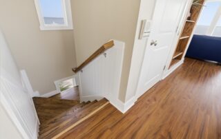 Wooden staircase with white railing leading down, adjacent to a bright room with hardwood floors. Southport rentals vibe with ocean view.