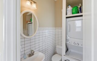 Compact bathroom with white tile, round mirror, sink, toilet, and stacked washer-dryer unit. Suitable for Southport rentals.