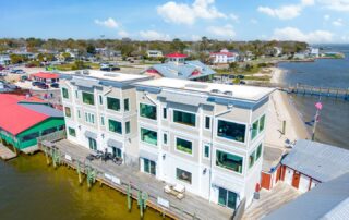 Waterfront building with wooden deck and large windows in a coastal area, ideal for Southport rentals. Nearby sandy beach, under blue skies.