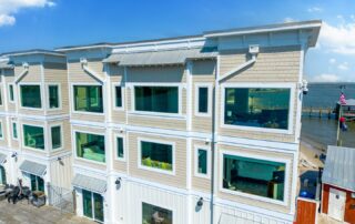 Three-story coastal building with large windows overlooking a dock and water, part of Southport rentals. Clear blue sky adds to the scenic view.