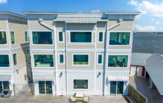 Three-story waterfront building with large windows and a wooden deck, associated with Southport rentals. Blue sky and calm water in the background.