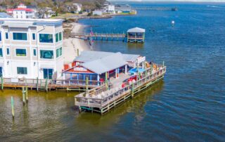 Waterfront dining spot by Southport rentals, featuring an outdoor deck, colorful umbrellas, and ocean views, adjacent to a sandy shoreline.