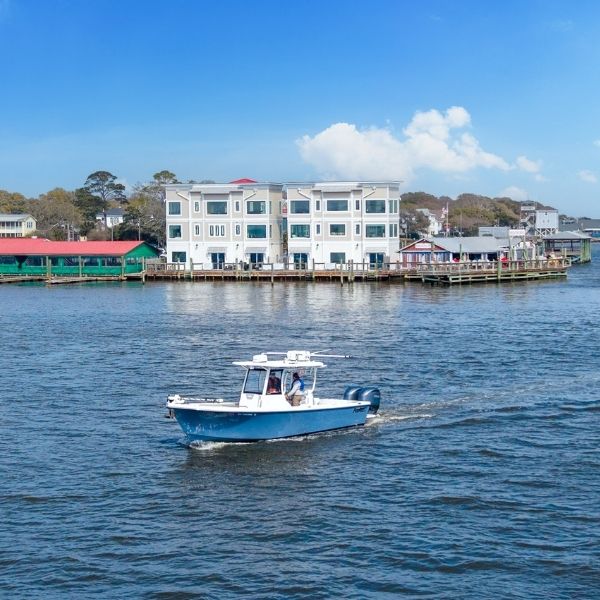 Blue boat cruising on calm water near waterfront buildings; Southport rentals visible in the background under a clear blue sky.